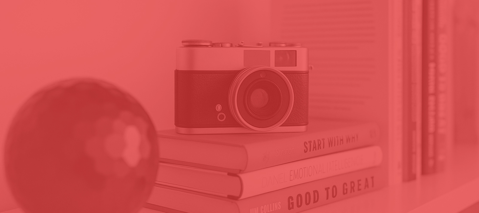 A vintage camera sits on a stack of books beside a spherical object, with additional books standing upright in the background. The entire image has a red color filter.