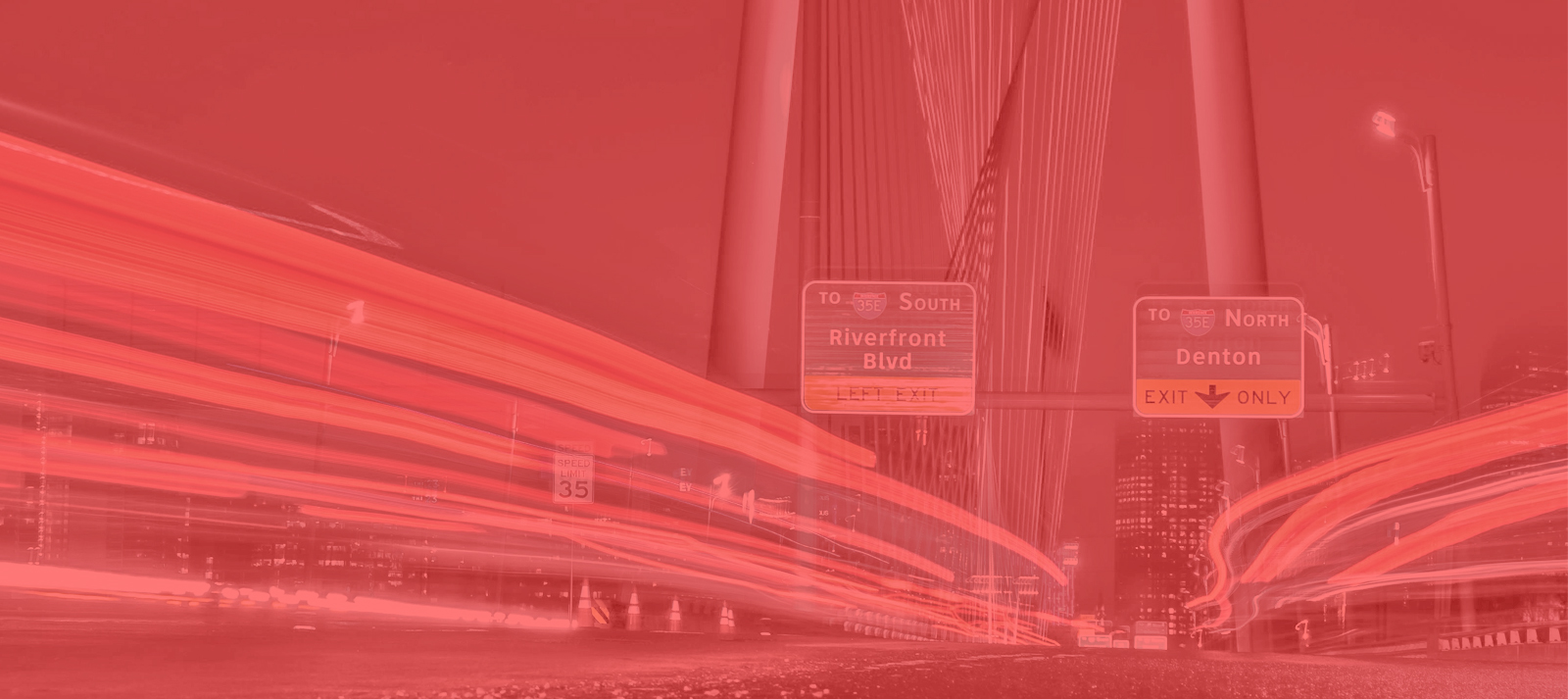 Long exposure photo of a city road at night with light trails from vehicles, bridge cables overhead, and highway signs pointing to Riverfront Blvd south and Denton north. The image has a red overlay.